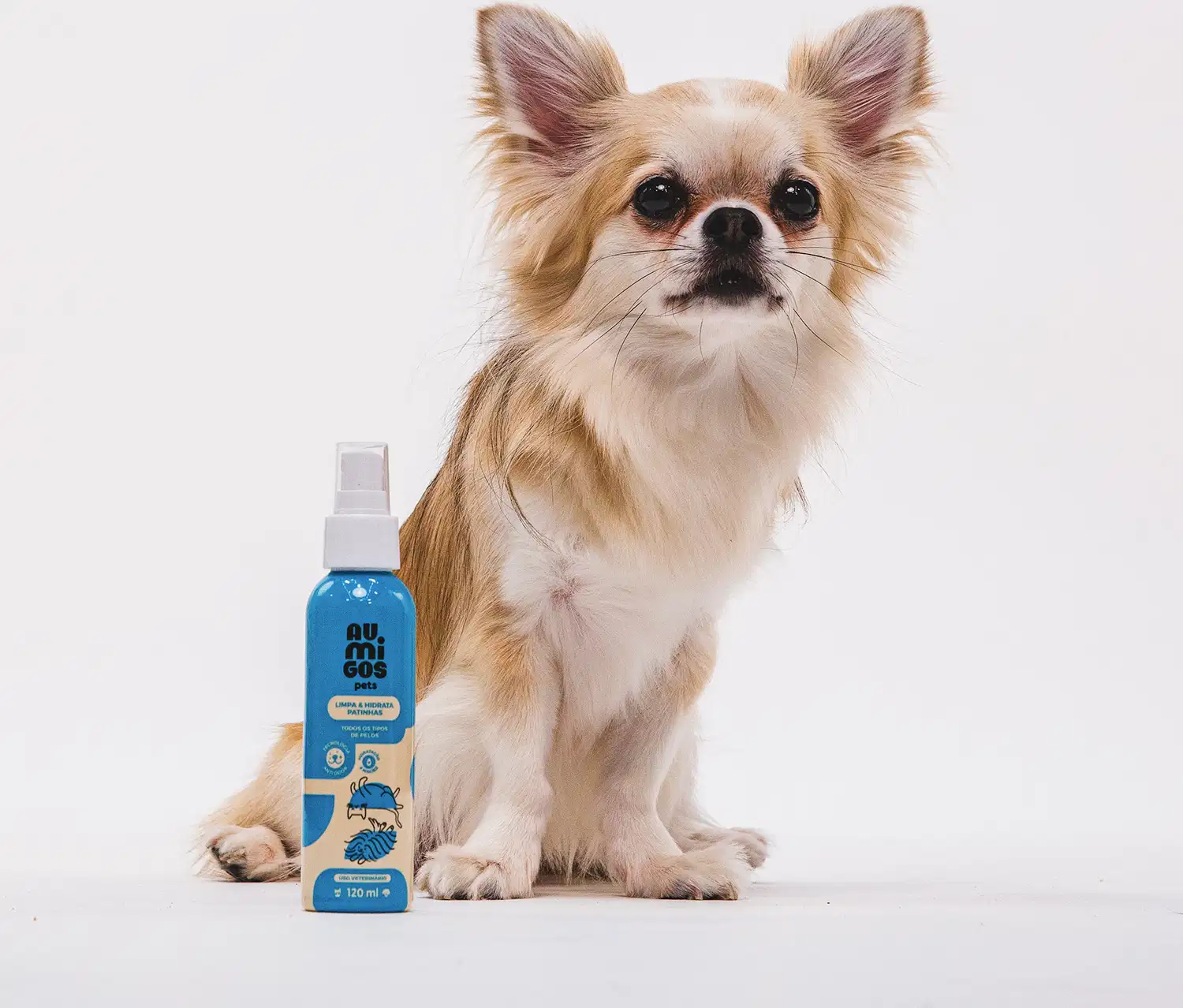 Frasco em tons de azul e rosa com tampa spray branca e letras pretas contendo 120ml de limpa e hidrata patinhas da marca Au.Migos. Ao lado da embalagem está um cachorrinho e o fundo é cinza claro.