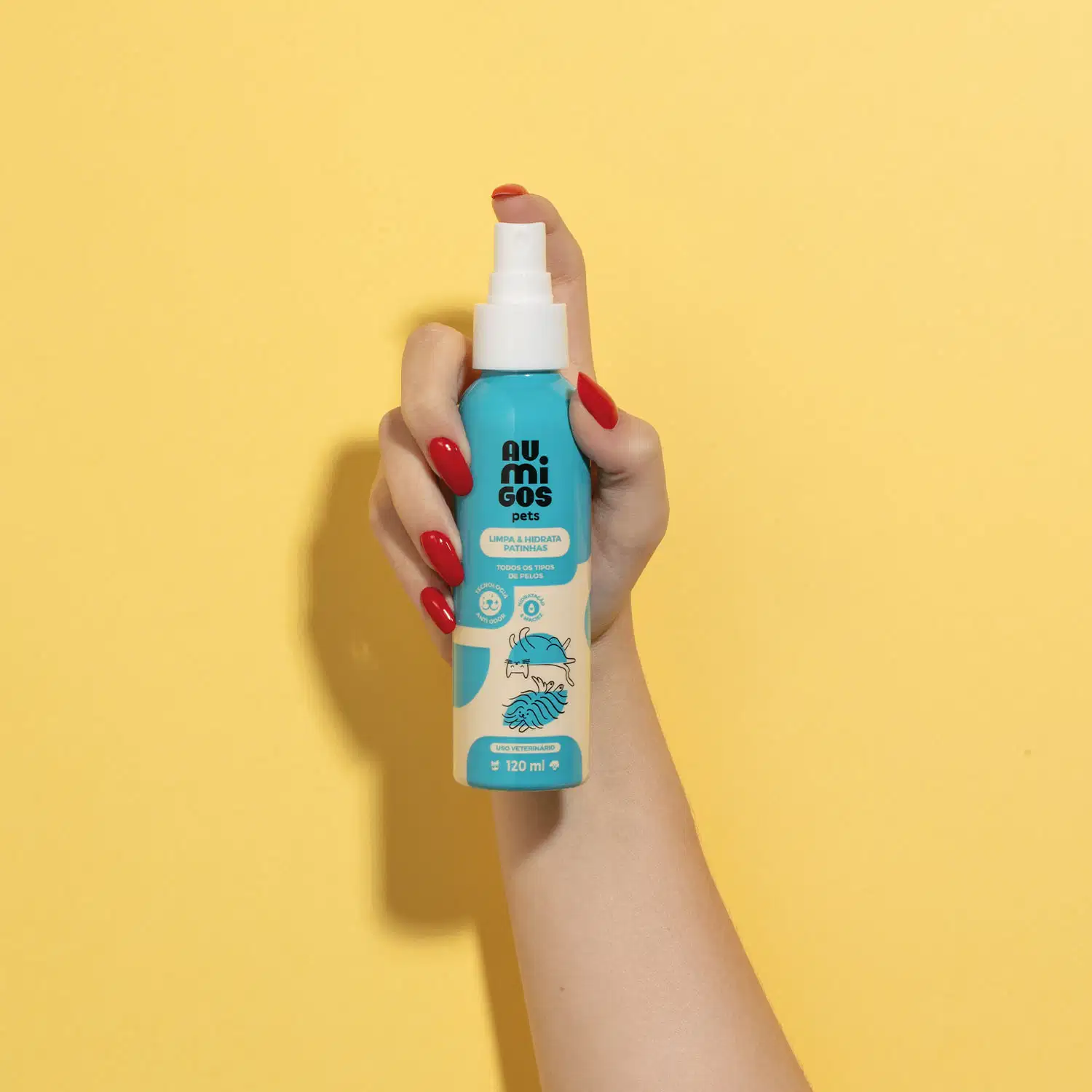 Mão segurando um Frasco em tons de azul e rosa com tampa spray branca e letras pretas contendo 120ml de limpa e hidrata patinhas da marca Au.Migos em fundo alaranjado.