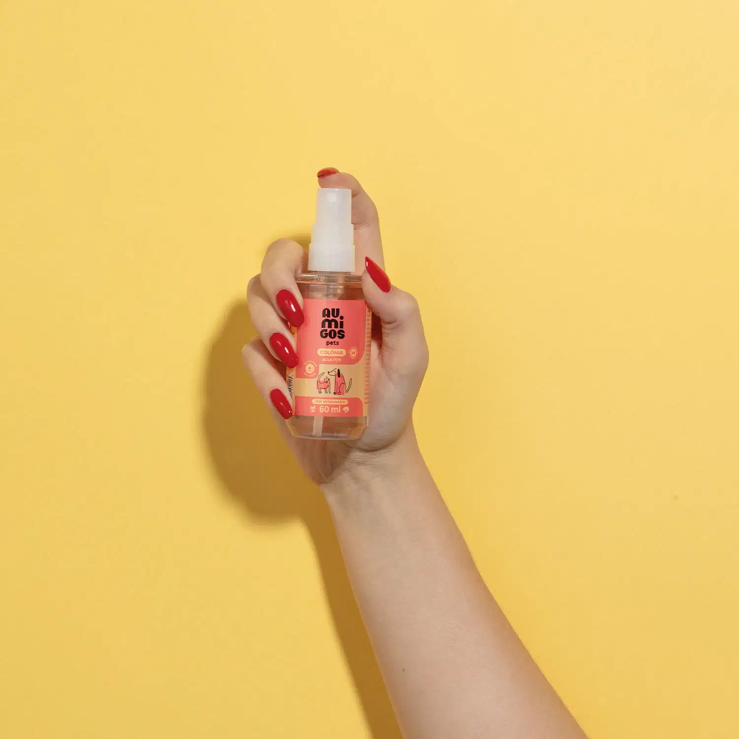 Mão segura um frasco com líquido claro, válvula spray branca e rótulo laranja e rosa com ilustrações de um gato e de um cachorro contendo 60ml de colônia adultos da marca Au.Migos Pets em fundo alaranjado.