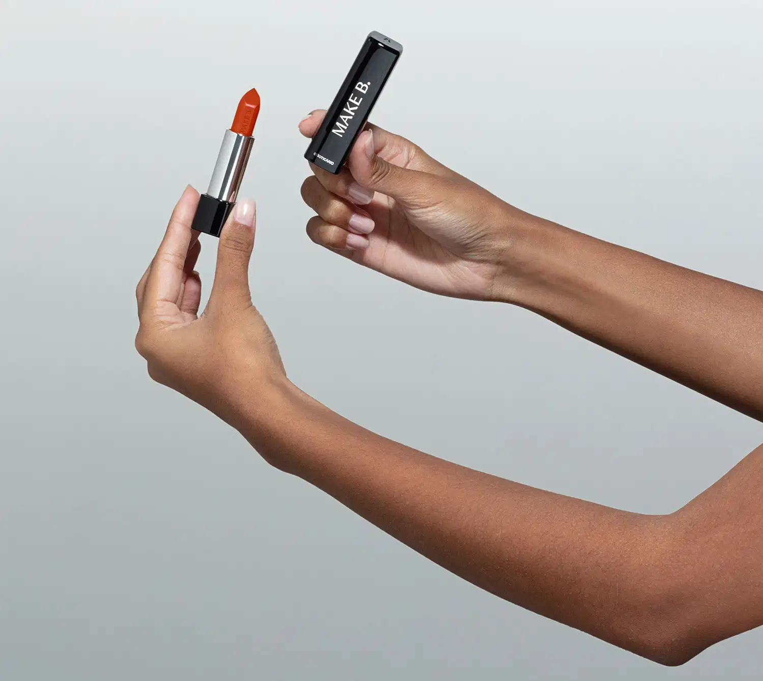 Uma mão feminina está segurando um Batom glossy na cor Coral da marca Make B. em embalagem prata e preta e a outra mão segura a tampa na cor preta com letras prateadas. O fundo da imagem é cinza.