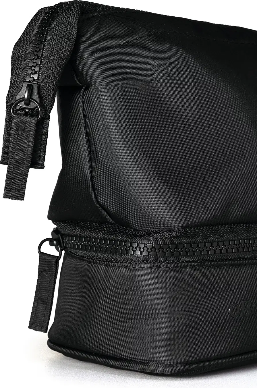 Close-up de Nécessaire masculina preta de nylon em formato triangular com foco na lateral que contém o puxador do zíper, com um puxador feito com pequena faixa retangular de nylon.