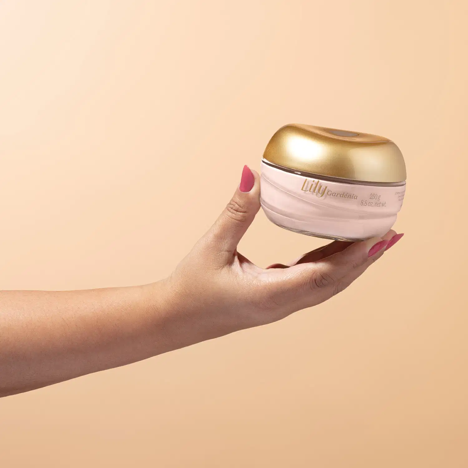 Mão feminina com unhas pintadas segurando um pote de vidro rosa claro com tampa dourada contendo creme hidratante corporal da marca Lily Gardênia em fundo bege.
