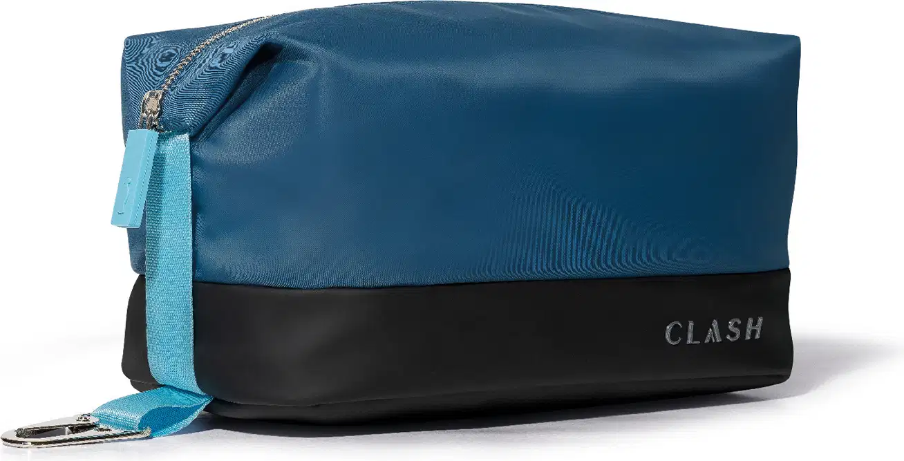 Bolsa de cosméticos azul-marinho com faixa preta na barra, com zíper e alça, exibindo a marca "CLASH" em letras grandes no canto inferior direito.  A vista lateral permite ver o detalhe em tecido azul mais claro do topo do zíper até a base do nécessaire.