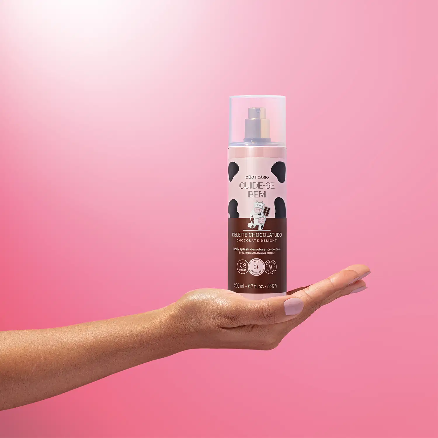 Mão feminina segurando um frasco de body splash Cuide-se Bem Deleite Chocolatudo em fundo rosa.
