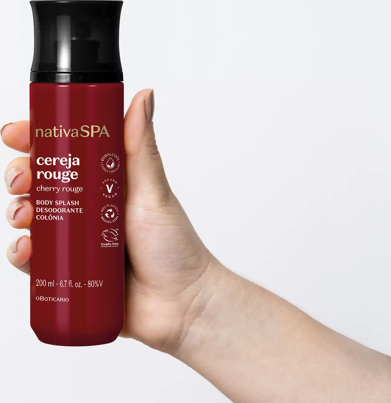 Mão feminina segurando um frasco vermelho escuro com tampa preta de body splash Nativa SPA Cereja Rouge em fundo azul claro.
