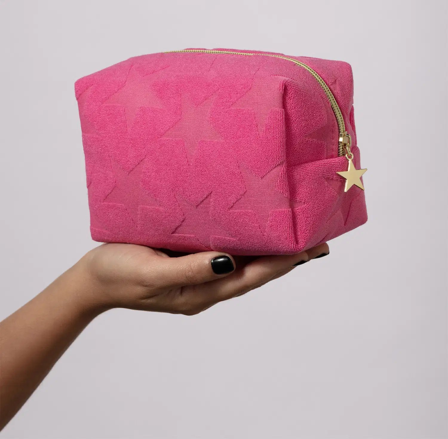 Mão segurando um nécessaire rosa macio com estampa de estrelas e fecho em zíper com pingente também de estrelas em fundo cinza claro.
