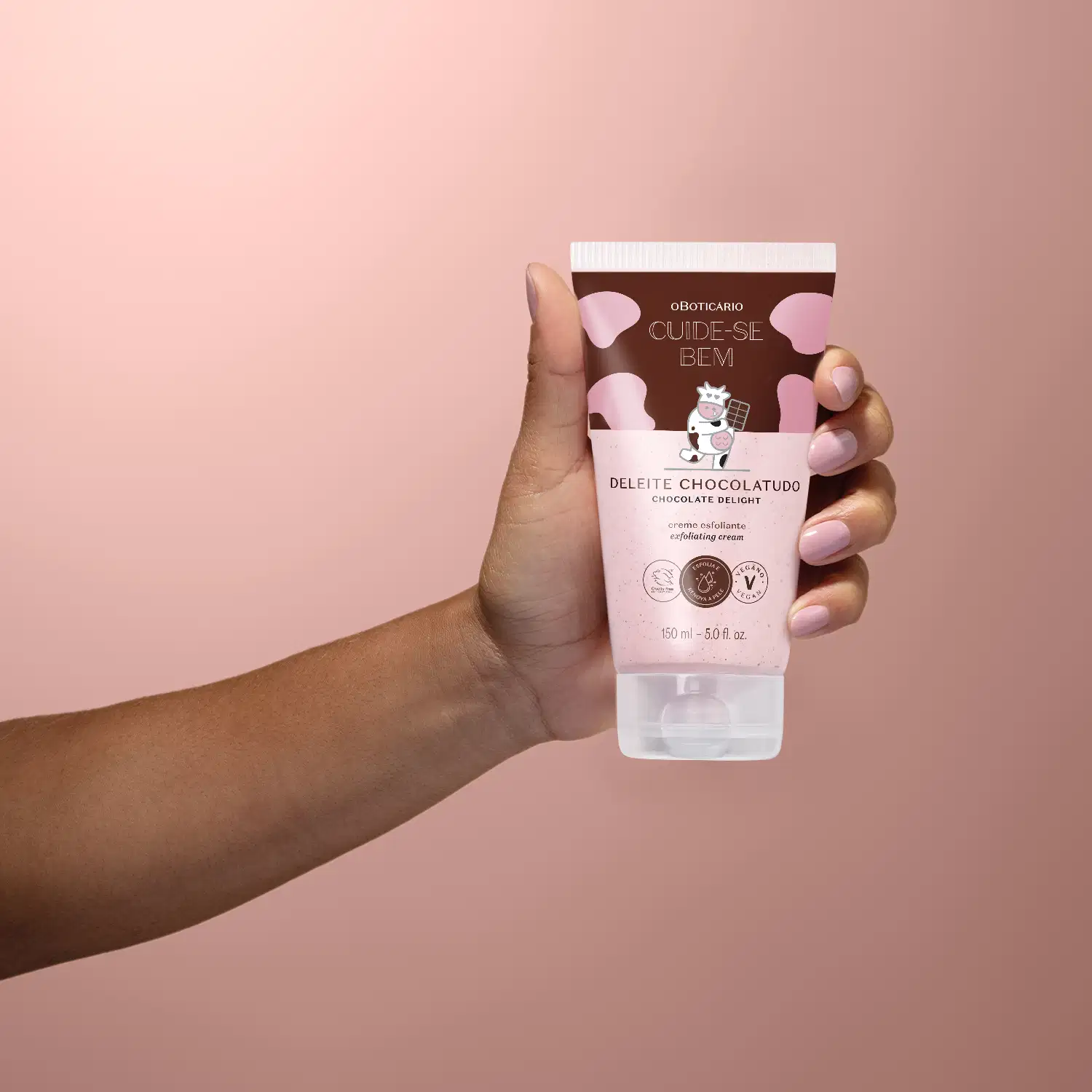 Mão feminina segurando um tubo de creme esfoliante corporal Cuide-se Bem Deleite Chocolatudo em fundo em tons de bege.