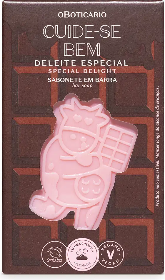 Sabonete em barra com vaca rosa estampada. Embalagem retangular marrom com texto "Cuide-se Bem Deleite Especial" e "Sabonete em Barra" em letras brancas. Selos indicando produto vegano e cruelty-free. Cores predominantes são marrom, rosa e branco.