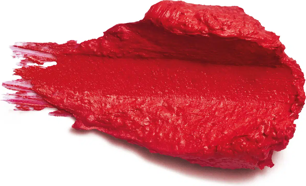 Amostra de batom de cor vermelha intensa. O produto tem uma textura espessa e cremosa, com uma superfície irregular e brilhante. Há vestígios do produto espalhados ao redor. 
