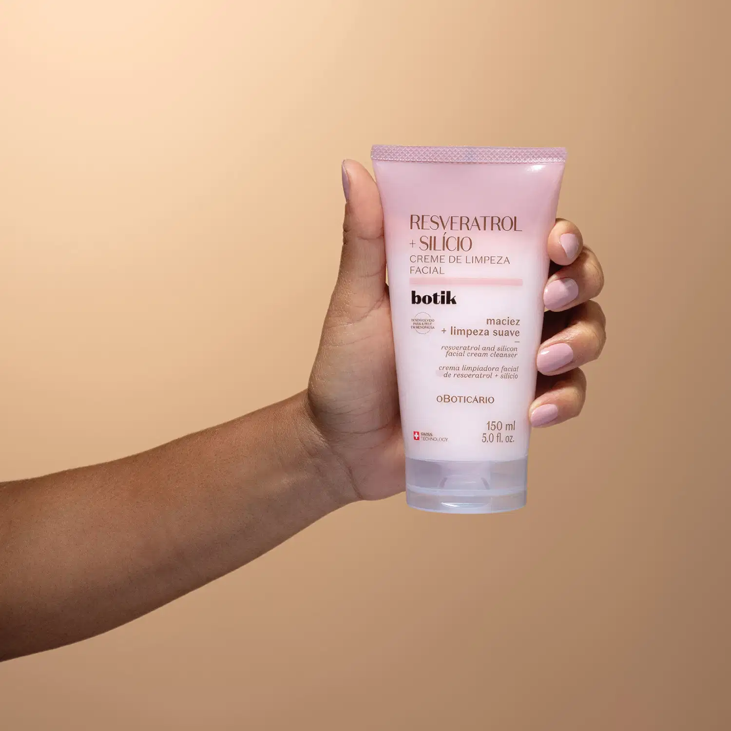 Mão segurando uma bisnaga com degradê de rosa e branco e letras marrons de creme de limpeza facial Botik  Resveratrol e Silício em fundo em tons de rosa.