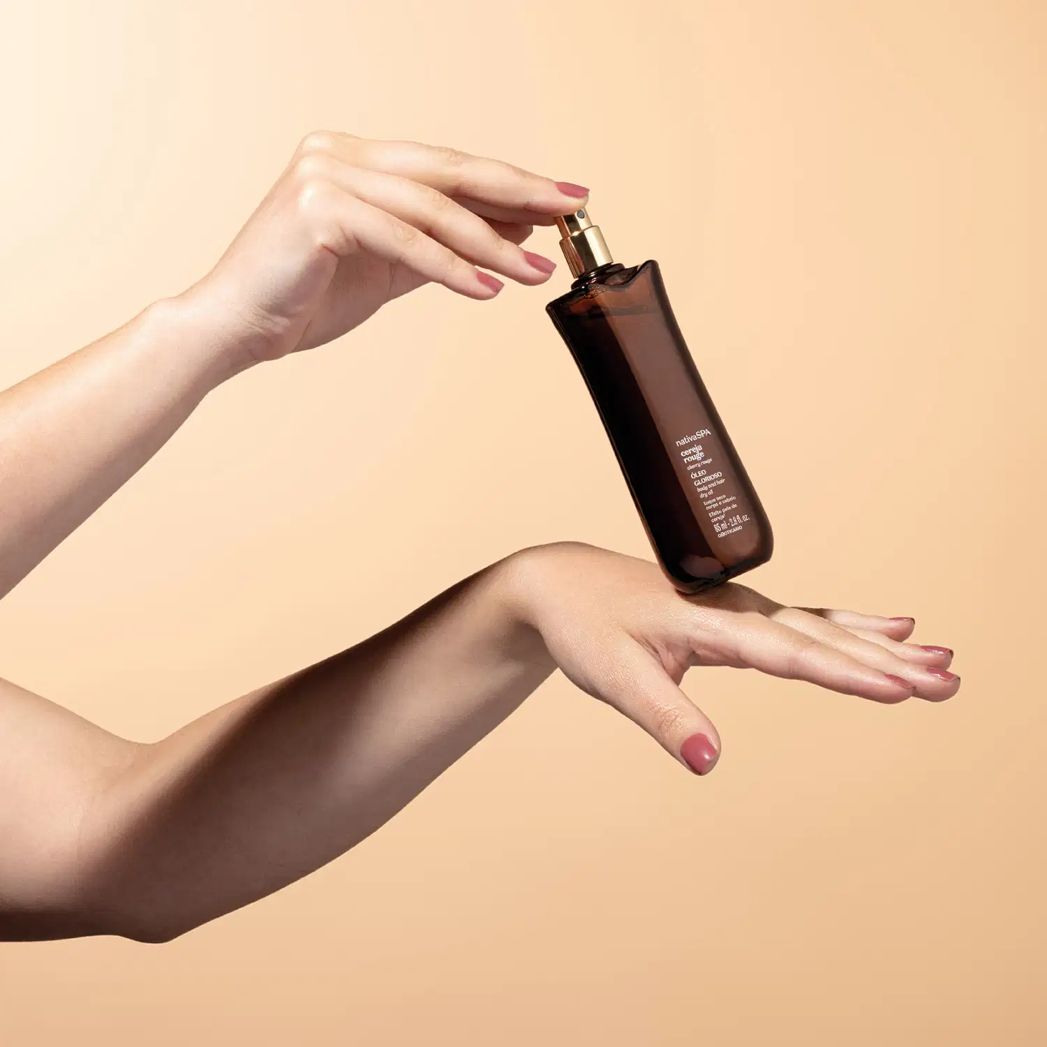 Mãos segurando um frasco de óleo glorioso para corpo e cabelos da marca Nativa SPA Cereja Rouge sobre fundo bege, com destaque para a embalagem escura e o gesto delicado.