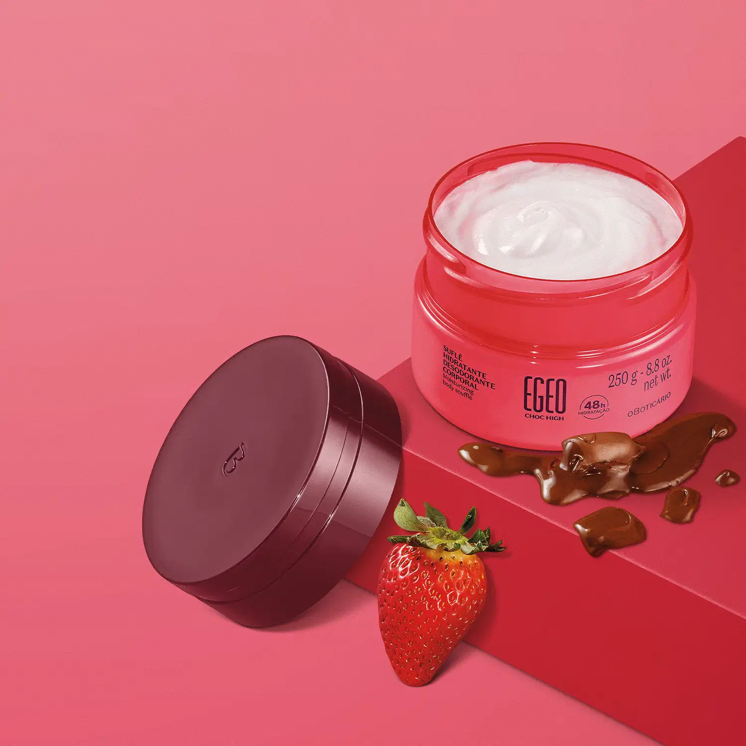 Pote aberto de Suflê Hidratante Desodorante Corporal da linha Egeo Choc High. A tampa vinho está apoiada em uma plataforma elevada pink, onde vemos o pote aberto do produto. Ao lado, vemos chocolate líquido e um morango sobre fundo rosa.