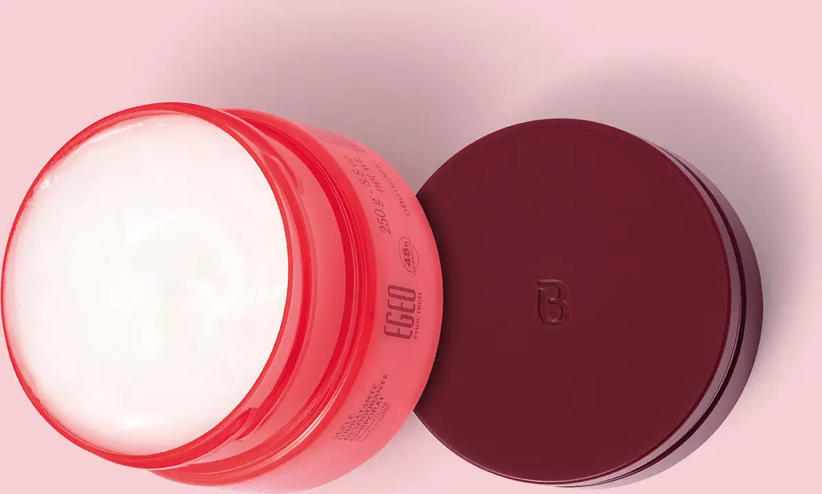 Pote aberto de Suflê Hidratante Desodorante Corporal da linha Egeo Choc High. A tampa vinho está sobre uma superfície rosa ao lado da embalagem, vistos de cima, o que permite ver o creme corporal branco dentro do pote rosa.