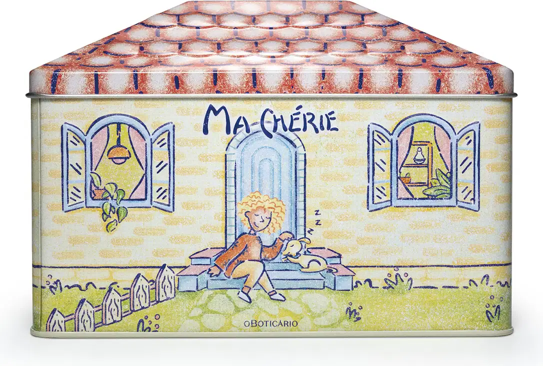 Lata retangular de metal decorada com ilustrações coloridas. A tampa apresenta um padrão de telhas vermelhas e brancas. Na parte frontal, há uma cena ilustrada com uma figura feminina loira sentada em um banco, segurando um livro, em frente a uma casa.