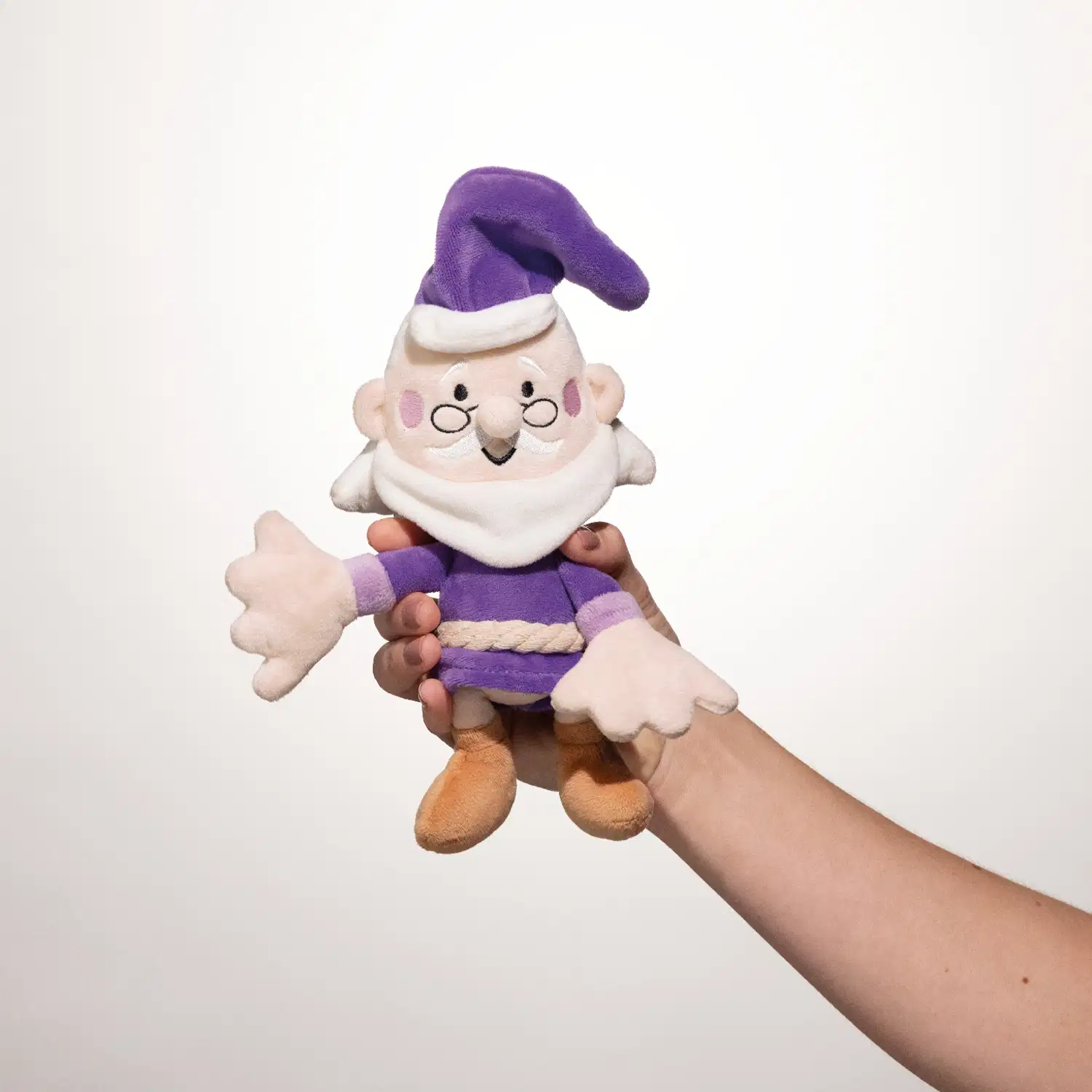Mão segurando um boneco de pelúcia, personagem da marca Dr. Botica, sendo um senhor idoso com barba branca, óculos e roupa roxa.