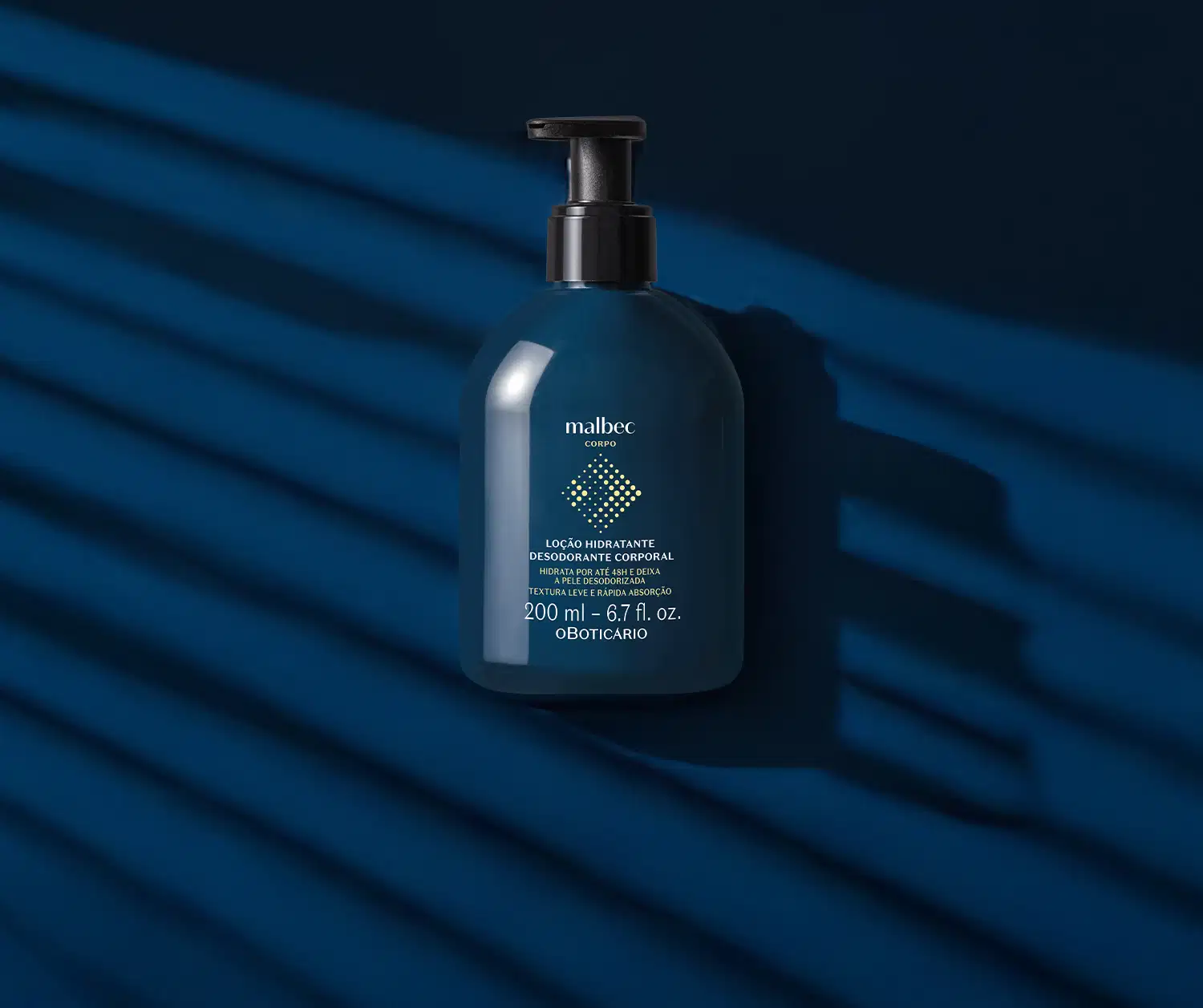 Frasco azul marinho com bomba dosadora preta, letras brancas e detalhes amarelos contendo 200ml de loção hidratante corporal Malbec Corpo em fundo azul escuro com muitas sombras formando listras no cenário.