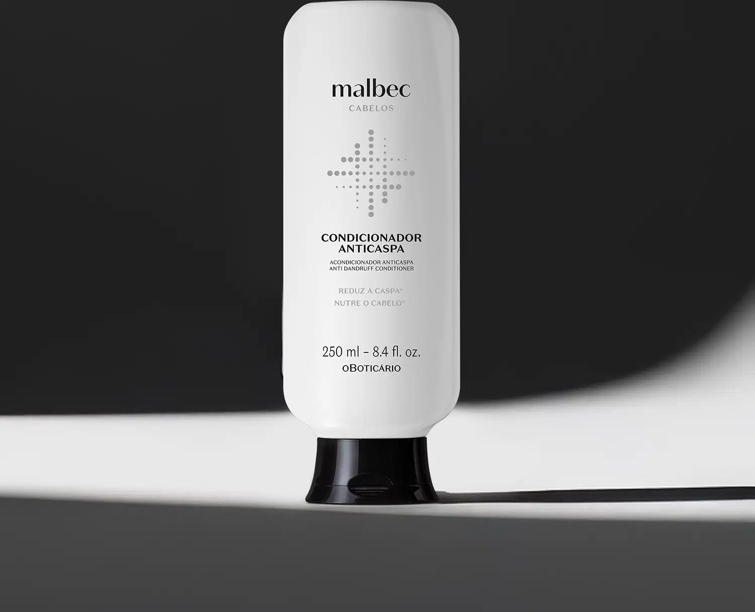 Imagem de frasco branco com tampa preta e rótulo com letras pretas da marca Malbec para cabelos, sobre superfície branca iluminada com luz cênica projetando uma sombra a direita do frasco sobre fundo preto.