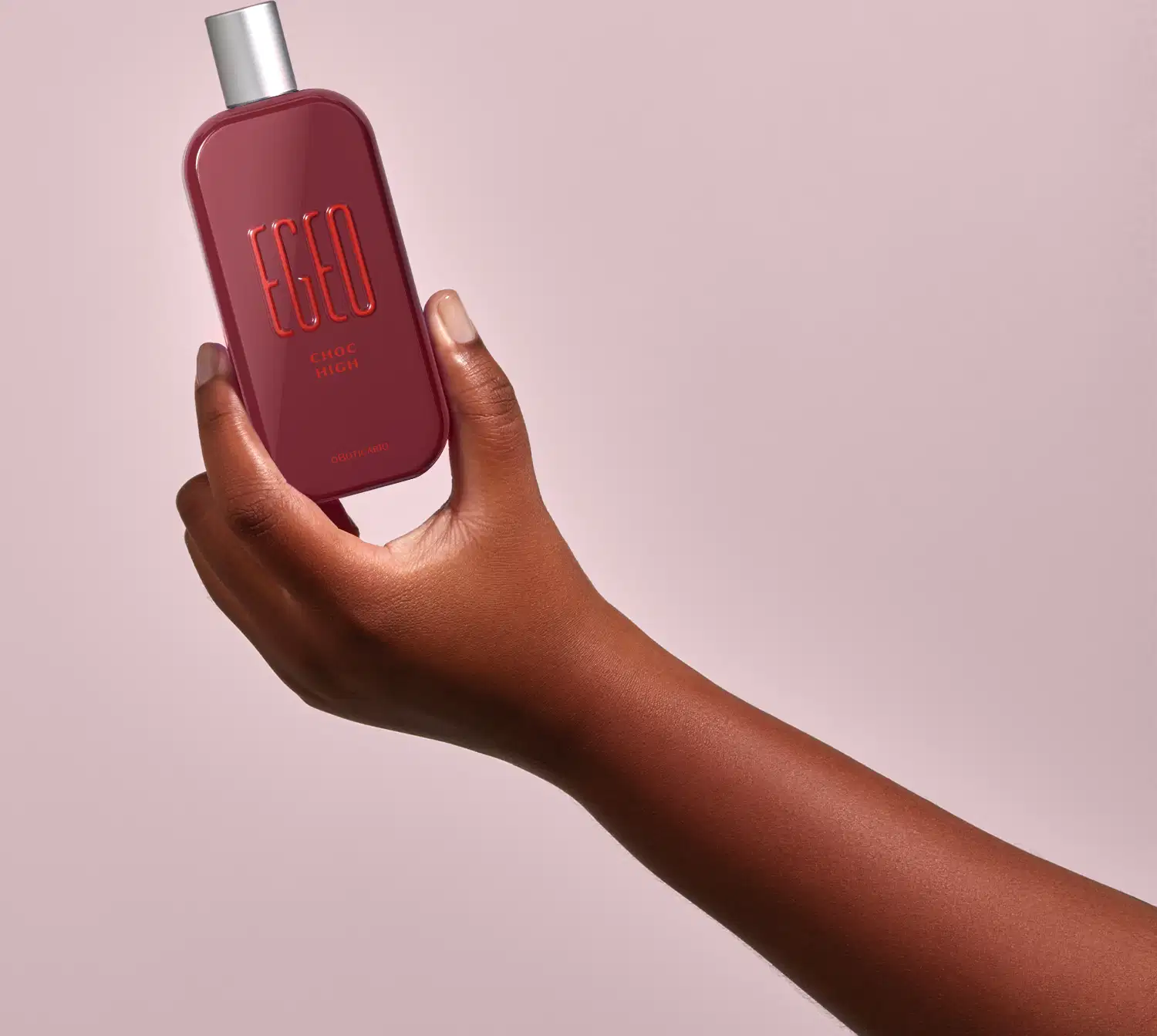 Frasco vermelho de desodorante colônia Egeo Choc High sendo segurado por mão feminina de pele negra contra fundo rosa claro.