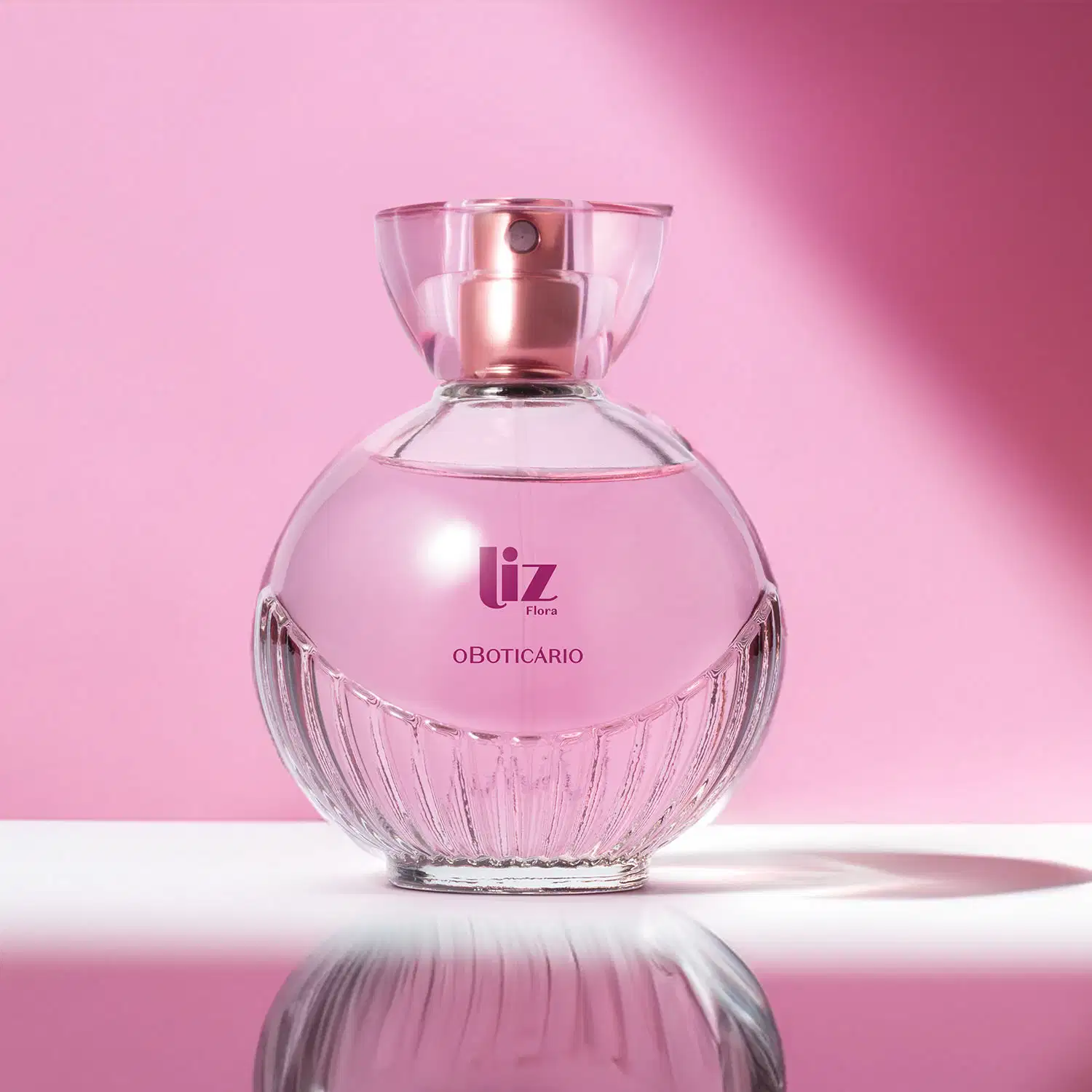 Vidro arredondado com líquido rosa, letras vinho, borrifador dourado e tampa transparente contendo desodorante colônia Liz Flora em fundo e superfície claros com algumas sombras.