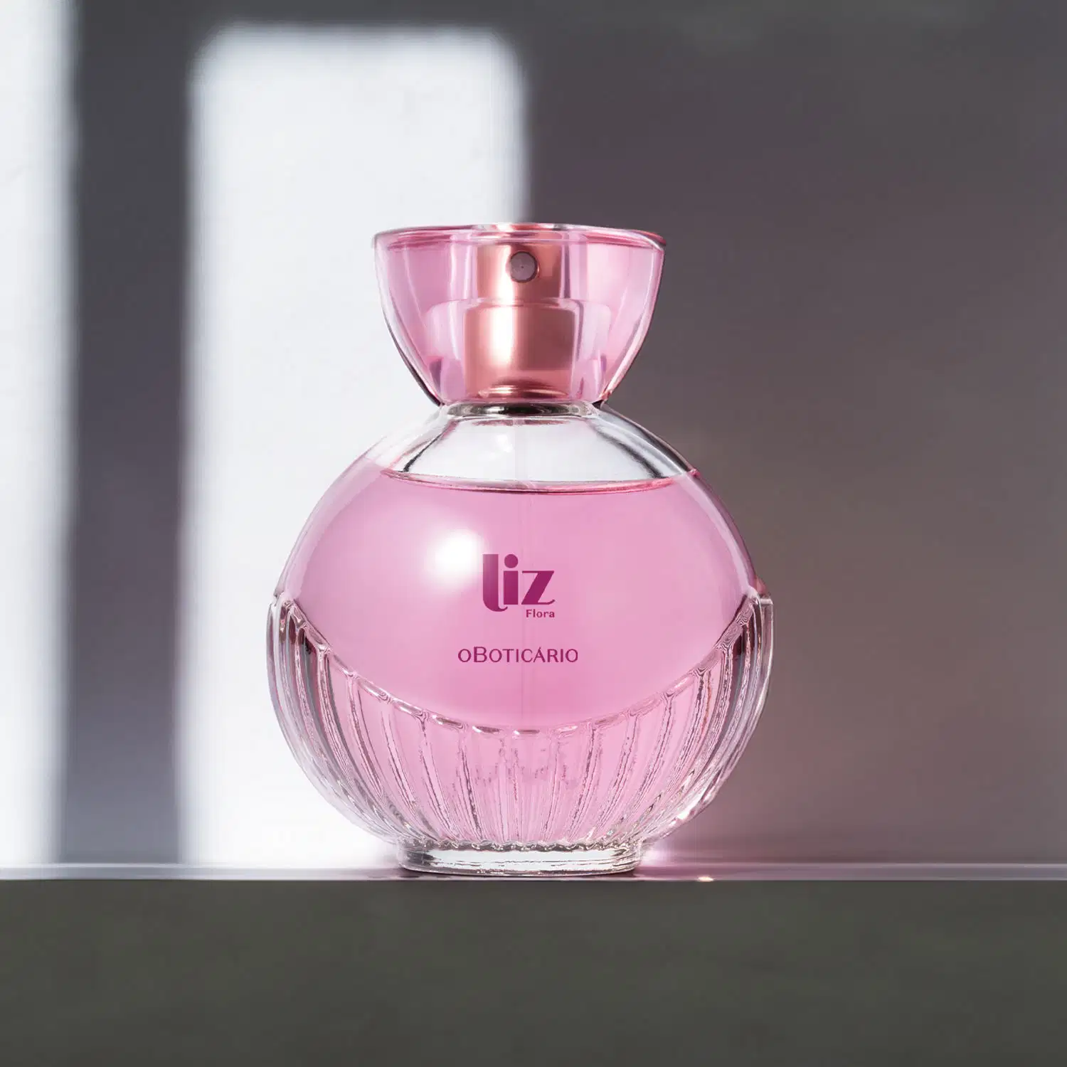 Vidro arredondado com líquido rosa, letras vinho, borrifador dourado e tampa transparente contendo desodorante colônia Liz Flora em fundo claro com muitas sombras.