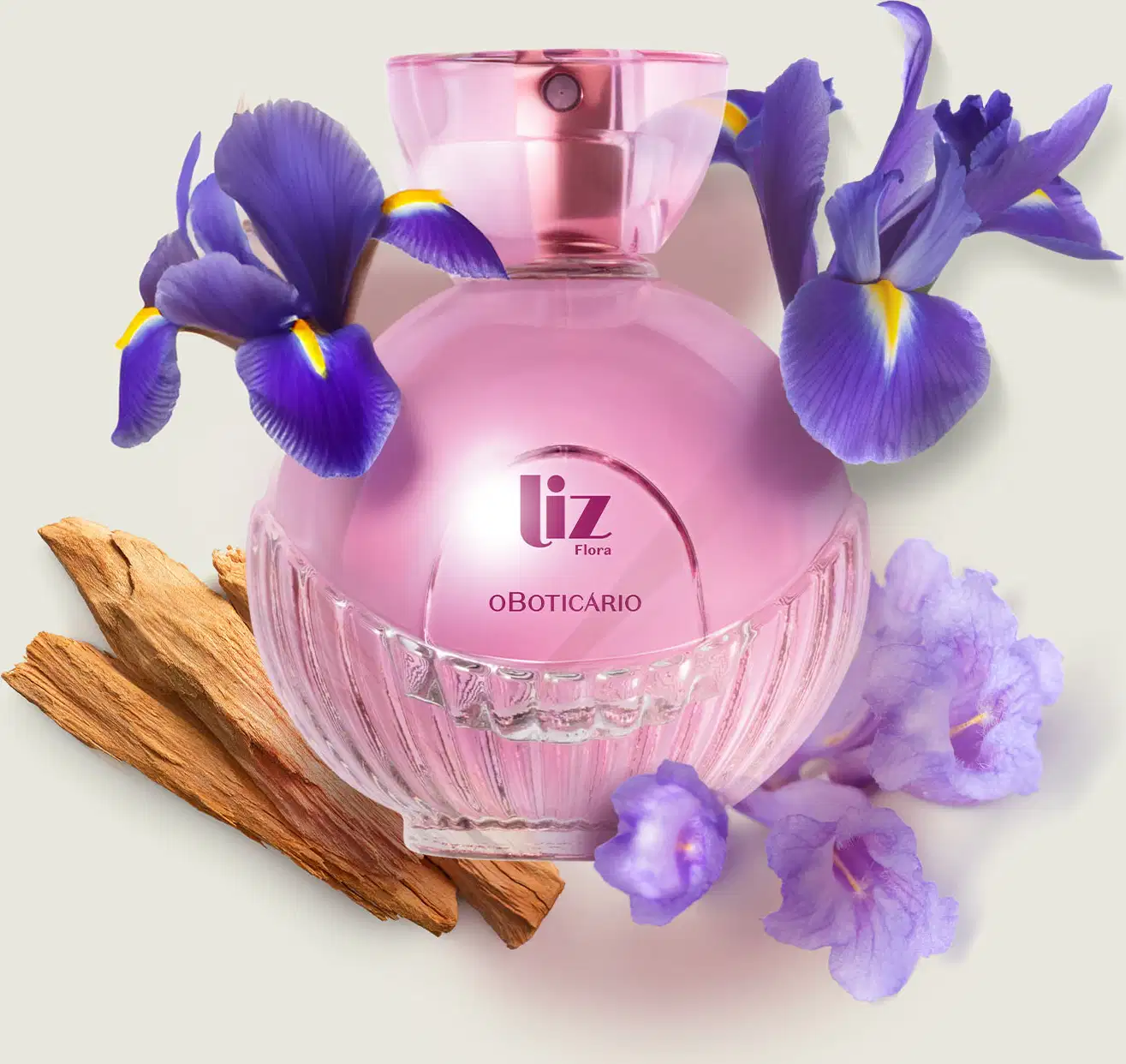 Vidro arredondado com líquido rosa, letras vinho, borrifador dourado e tampa transparente contendo desodorante colônia Liz Flora rodeado por flores de íris violetas e pedaços de madeira em superfície clara.
