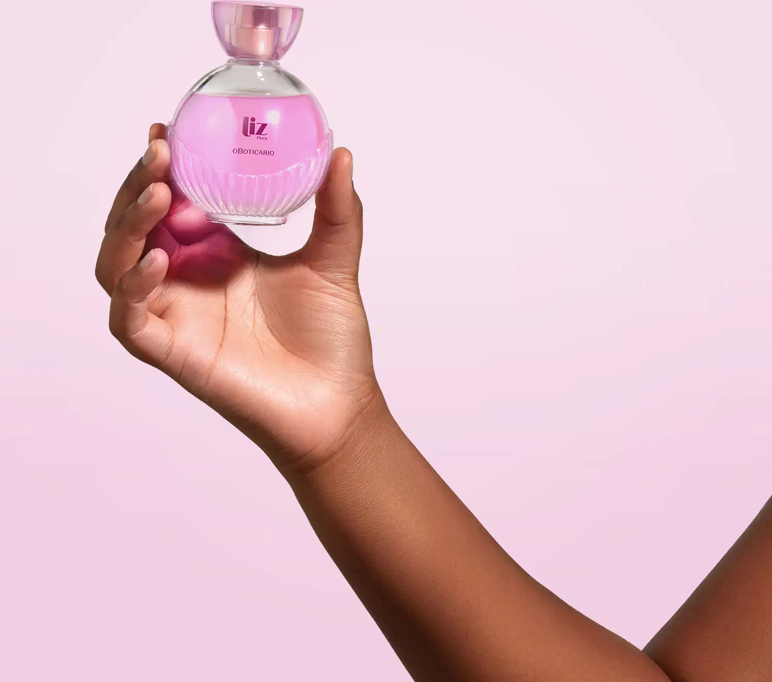 Mão feminina segurando um vidro arredondado com líquido rosa, letras vinho, borrifador dourado e tampa transparente contendo desodorante colônia Liz Flora em fundo lilás claro.