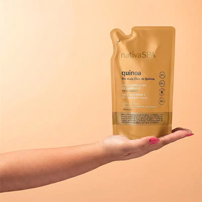 Imagem de um braço estendido e a mão segurando uma embalagem refiil de loção corporal da marca Nativa SPA Quinoa, com sachê de plástico flexível na cor dourada, rótulo com texto e informações sobre o produto em letras brancas e pretas.