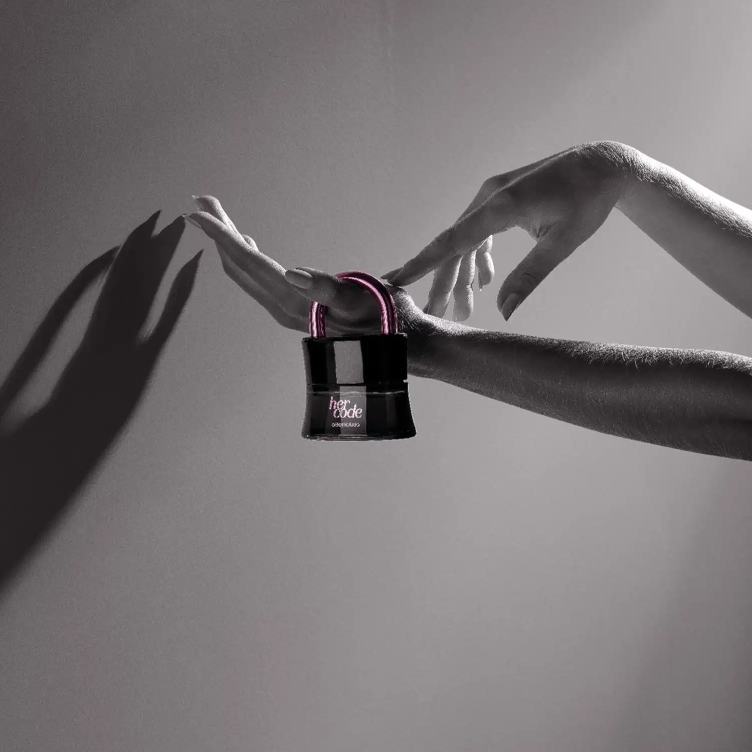 Mãos femininas segurando um vidro preto em formato de cadeado com arco e letras na cor rosa metalizado de Eau de Parfum Her Code Touch em fundo cinza escuro com sombras.