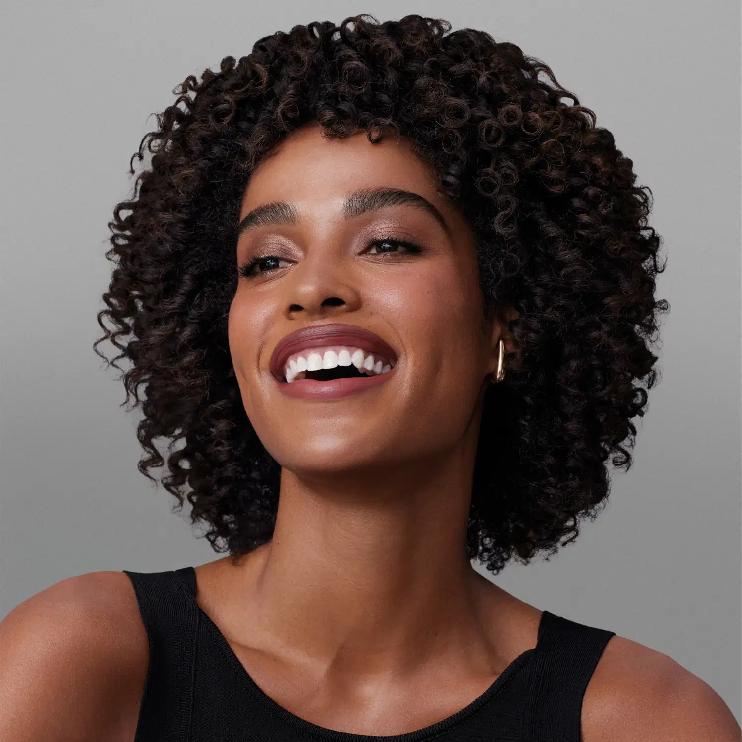 Ambiente de beleza com modelo sorridente, cabelos cacheados escuros e pele negra. Cenário neutro de fundo cinza sugere uso de produto para cuidados capilares da marca Match.