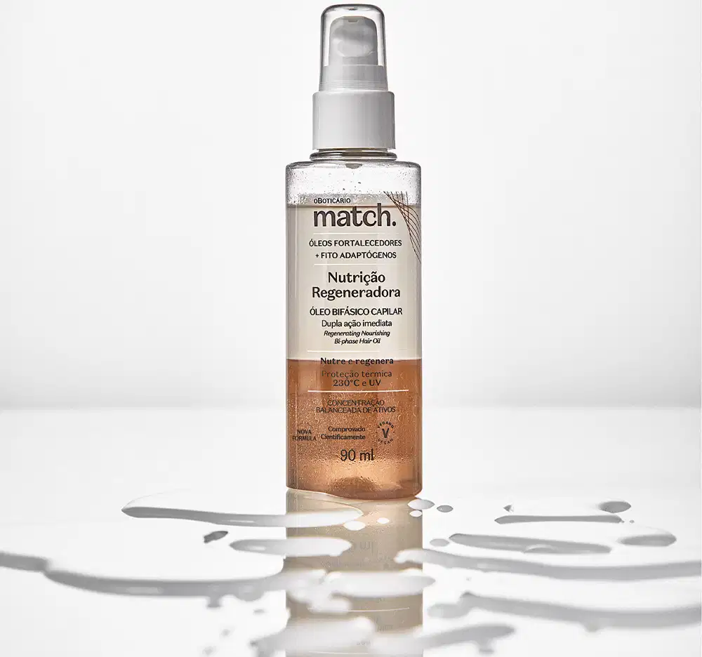 Imagem do um frasco transparente do Óleo Bifásico Capilar Match Nutrição Regeneradora em superfície transparente e molhada. A embalagem possui válvula spray e rótulo laranja com letras pretas. Há respingos de água por todo o frasco.