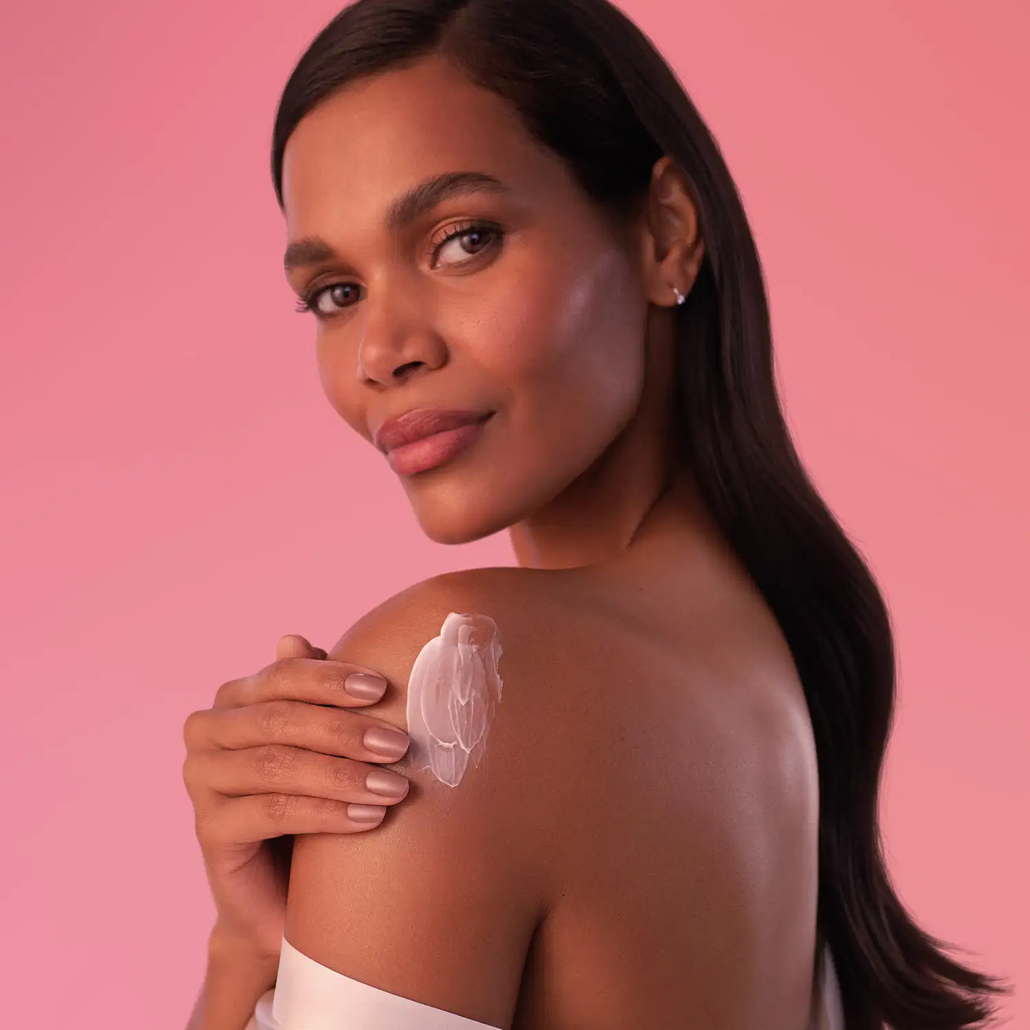 Mulher de pele negra com cabelo longo e liso de perfil passa creme hidratante sobre o ombro em um fundo rosa sólido, com expressão séria e olhar direto para a câmera.