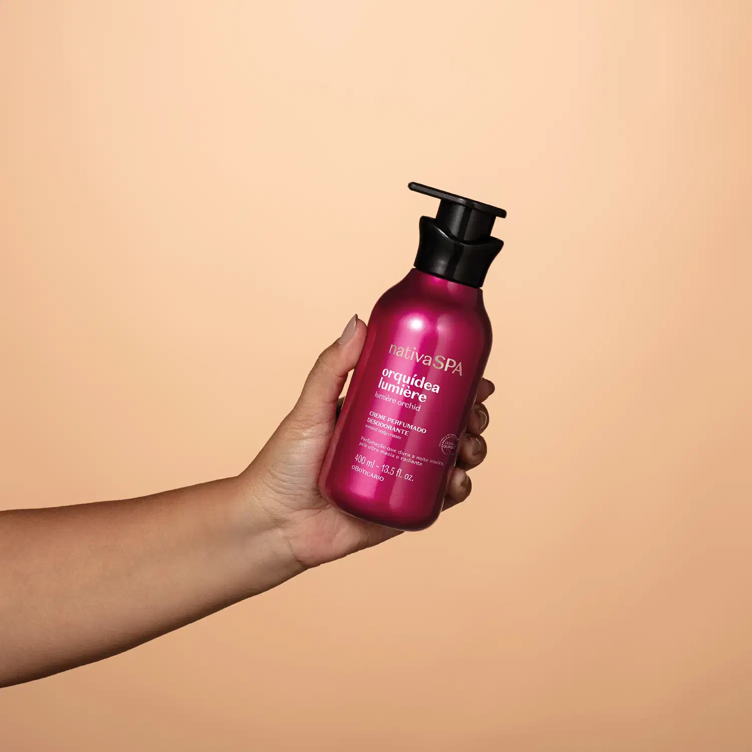 Embalagem pump rosa brilhante com tampa preta de item de corpo e banho da marca Nativa SPA Orquídea Lumiere sendo segurada por mão feminina em fundo laranja claro.