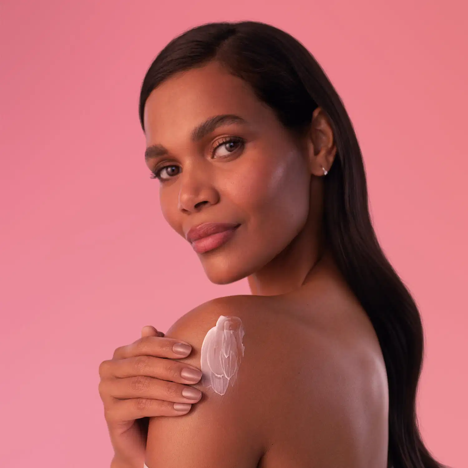 Mulher de pele negra com cabelo longo e liso de perfil passa creme hidratante sobre o ombro em um fundo rosa sólido, com expressão séria e olhar direto para a câmera.