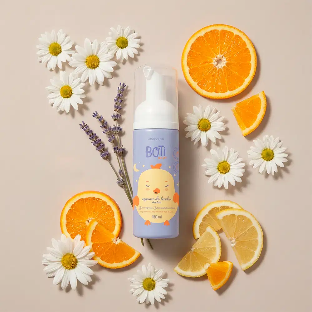A embalagem azul da espuma de banho Boti Baby é apresentado em um ambiente natural, com flores de camomila brancas e folhas de lavanda, além de fatias de laranja e limão, em um fundo bege claro, sugerindo um contexto de relaxamento e cuidados com a pele.