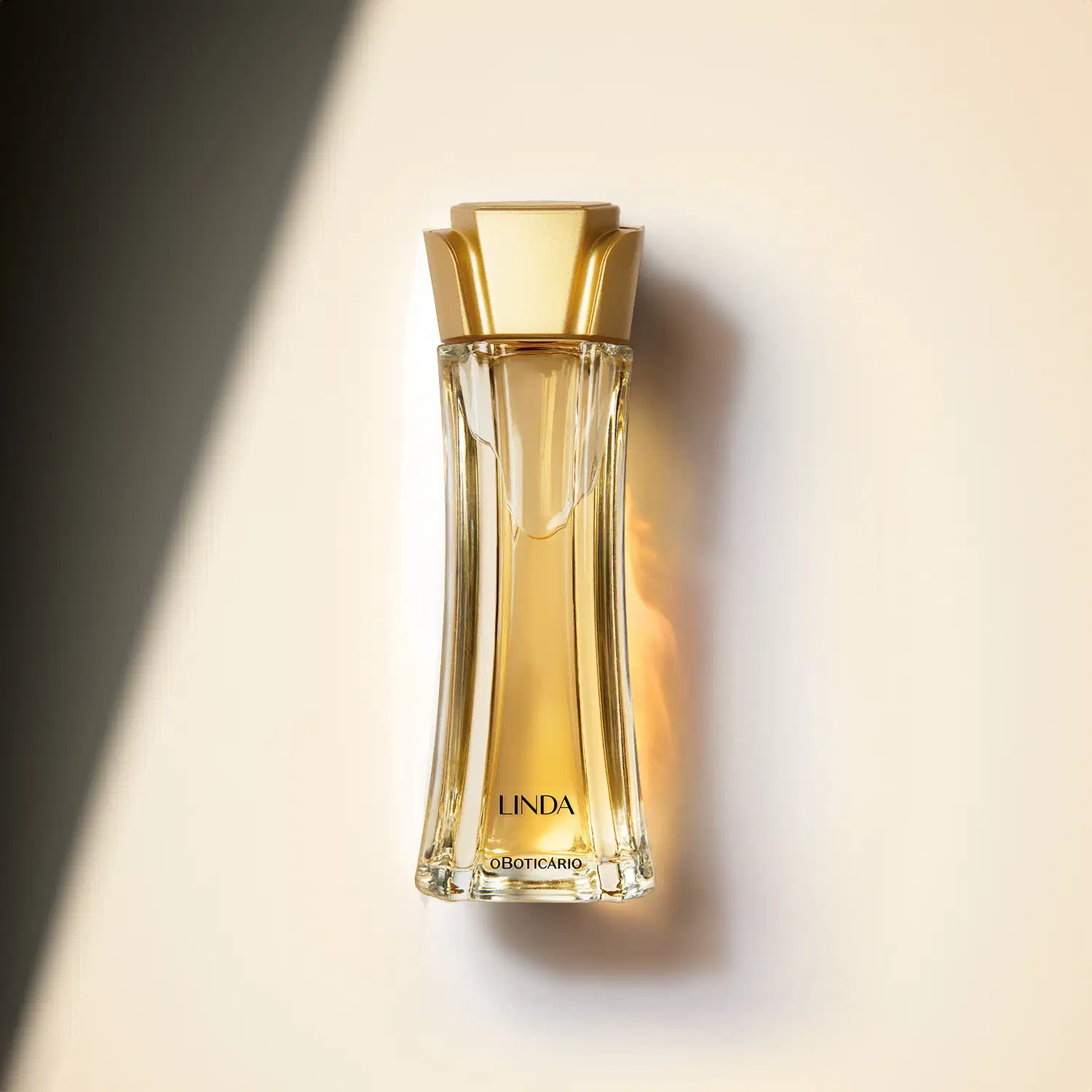 Vidro alongado com líquido bege e tampa dourada em forma de flor de desodorante colônia da marca Linda em um fundo claro com sombras.