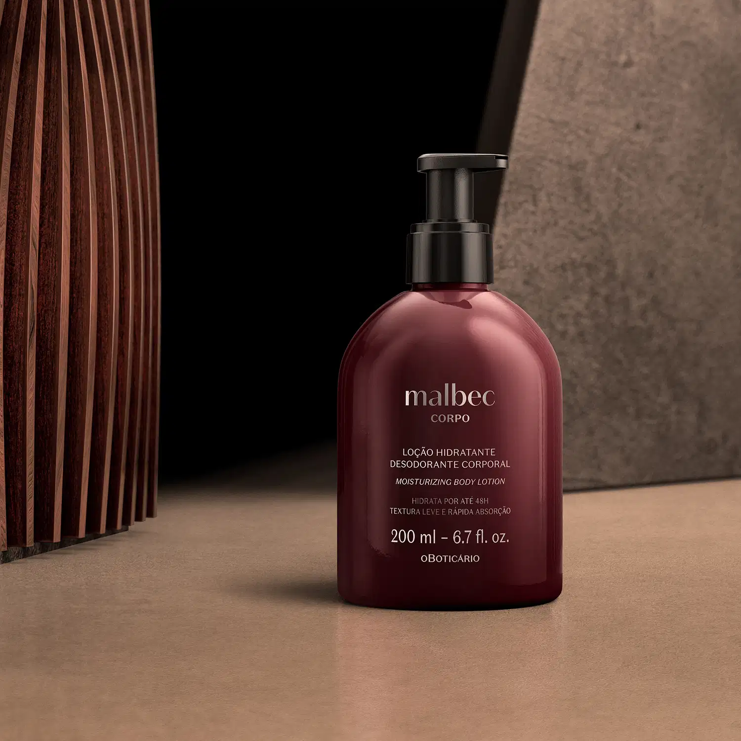 Frasco marrom de hidratante desodorante com a marca Malbec gravada em branco na embalagem em ambiente interno que sugere decoração em madeira ripada, pedra cinza e superfície bege.