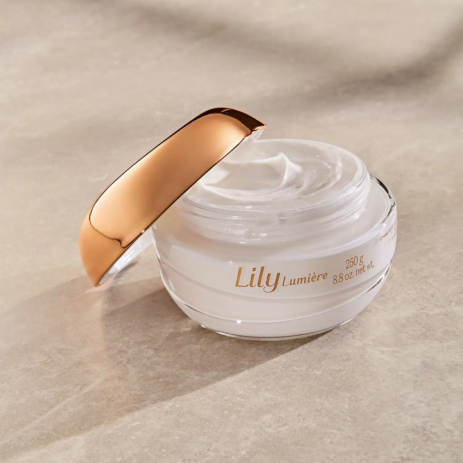 Pote de creme acetinado hidratante corporal Lily Lumière branco com tampa dourada aberto sobre superfície bege clara com sombra, mostrando textura cremosa do produto.