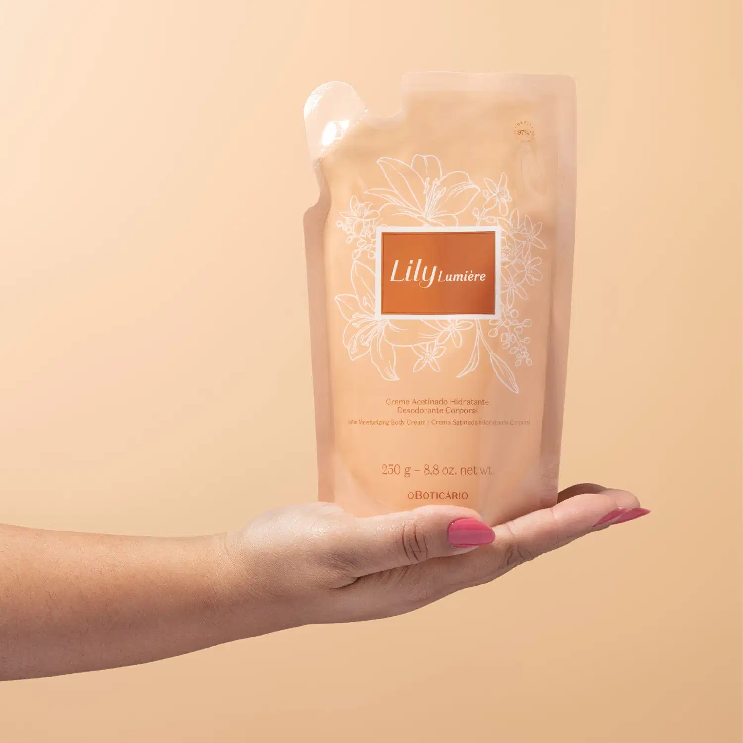 Mão feminina segurando um Refil de creme acetinado hidratante corporal da marca Lily Lumière em sachê de 250g na cor rosa com rótulo bege escuro, detalhes e letras douradas, sobre fundo rosa.