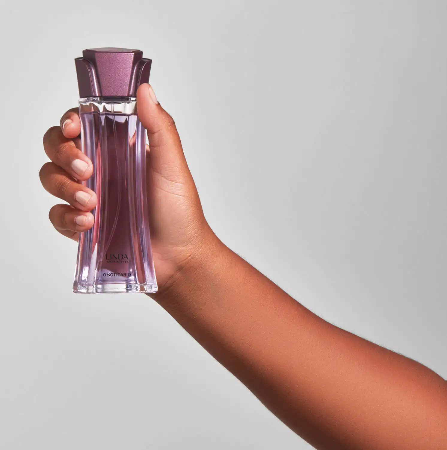 Mão segurando um vidro longo e transparente com líquido lilás, tampa roxa em formato de flor e letras roxas de desodorante colônia Linda Irresistível em fundo lilás.