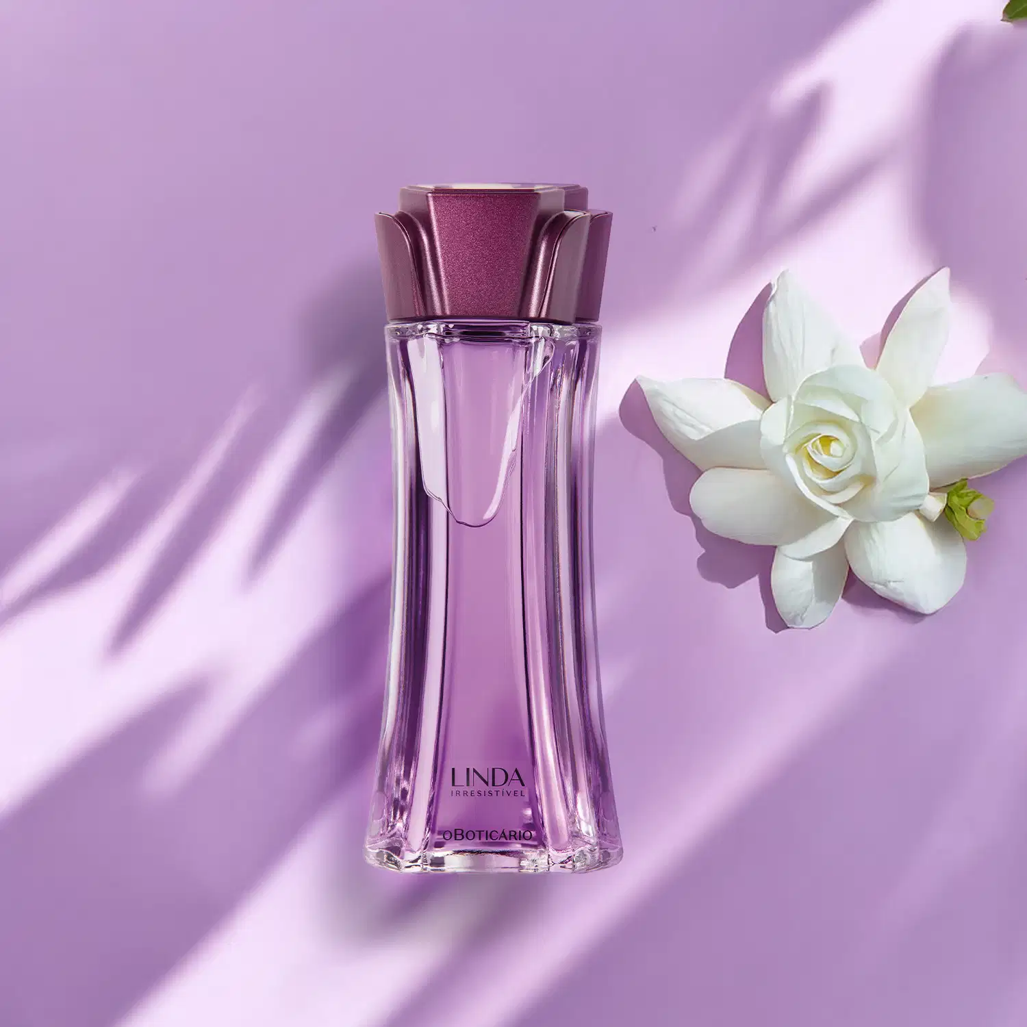 Vidro longo e transparente com líquido lilás, tampa roxa em formato de flor e letras roxas de desodorante colônia Linda Irresistível com camélia branca ao lado em fundo lilás com sombras.