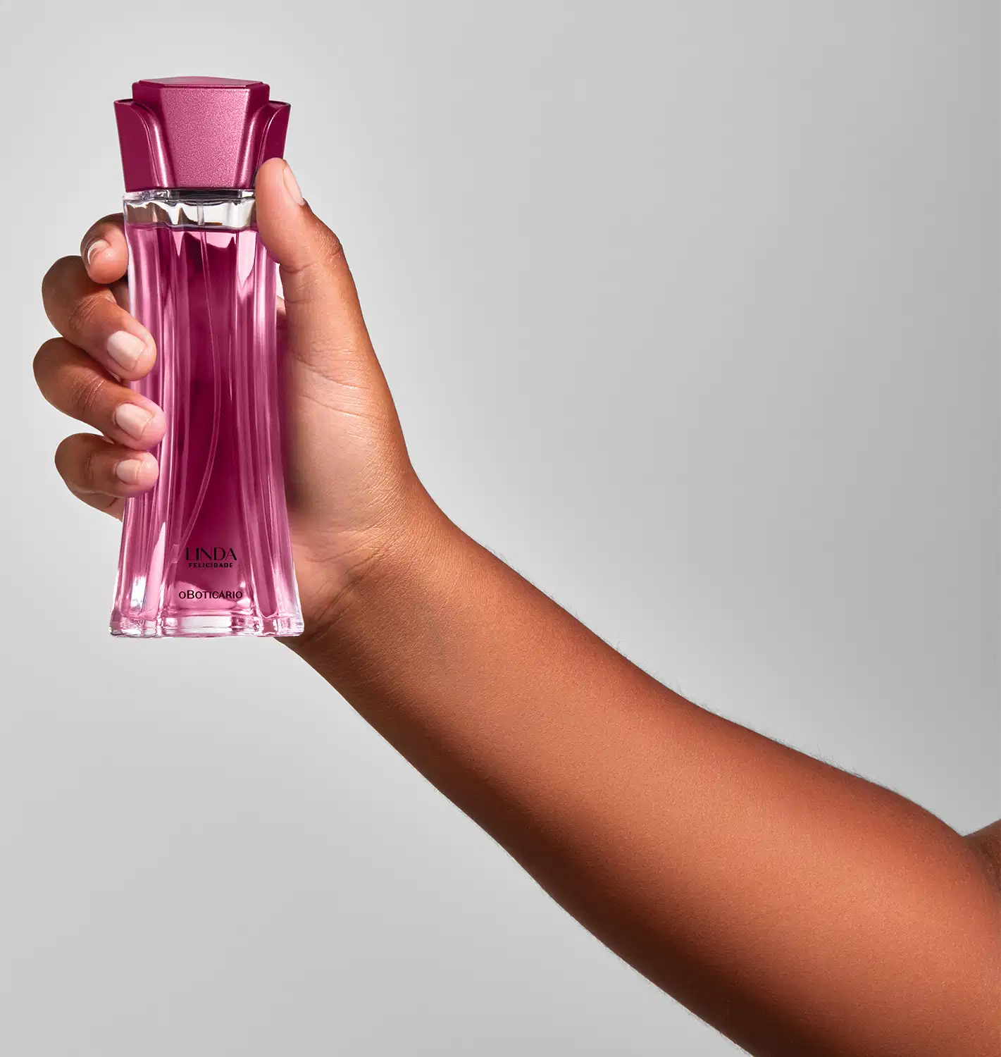 Mão feminina segurando um frasco de vidro com líquido rosa e tampa rosa em formato de flor contendo 100ml de desodorante colônia Linda Felicidade em fundo cinza.