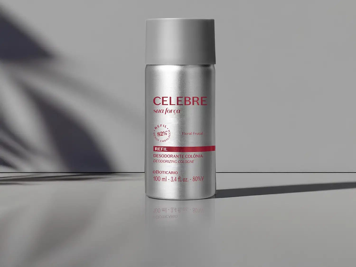 Refil de desodorante colônia Celebre Sua Força Feminino em frasco cilíndrico prateado com tampa cinza e letras vinho sobre superfície cinza com sombra de folha ao fundo, em ambiente com iluminação natural lateral.