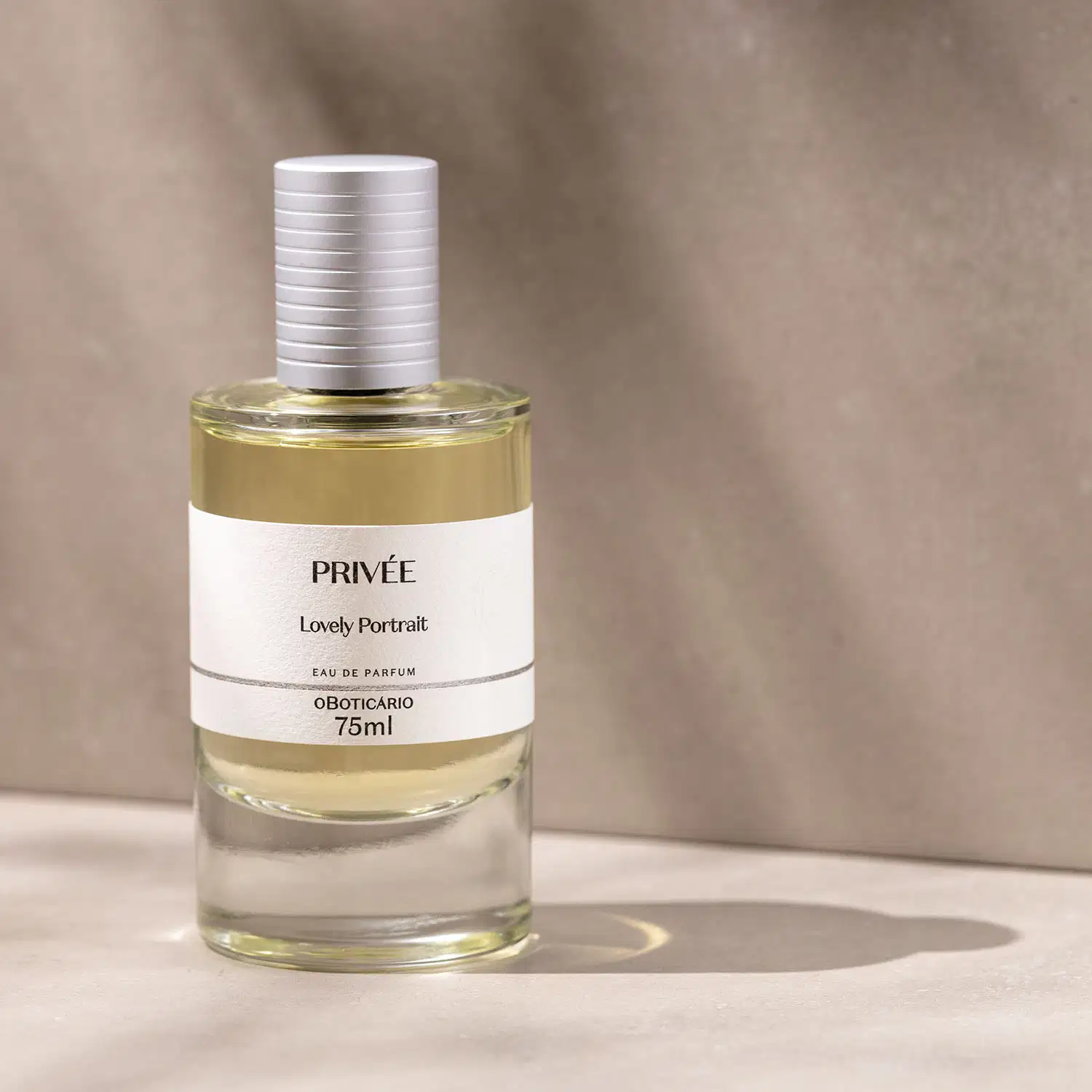 Vidro cilíndrico transparente com líquido bege, tampa prata e rótulo branco com letras pretas de Eau de Parfum Privée Lovely Portrait em superfície clara com sombras e fundo rosado marmorizado.