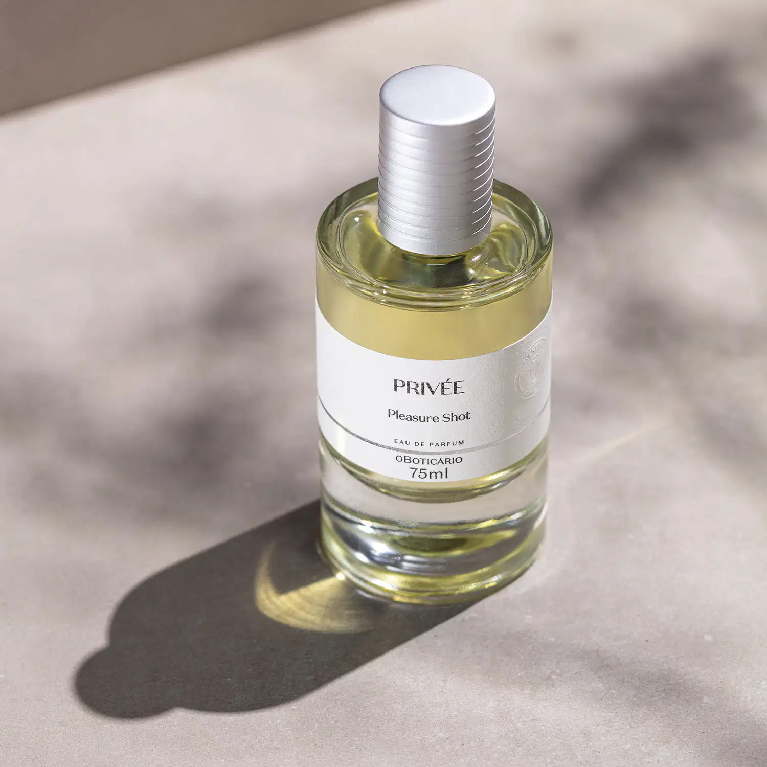 Vidro cilíndrico transparente com líquido amarelado, tampa prata e rótulo branco com letras pretas de Eau de Parfum Privée Pleasure Shot em um cenário claro com sombras.