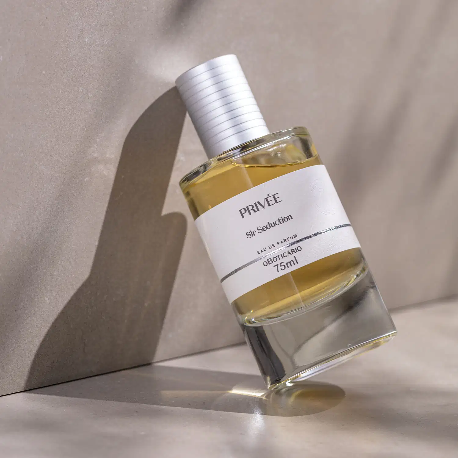 Vidro cilíndrico transparente com líquido alaranjado, tampa prata e rótulo branco com letras pretas de Eau de Parfum Privée Sir Seduction encostrado em um fundo marmorizado bege e superfície da mesma cor.