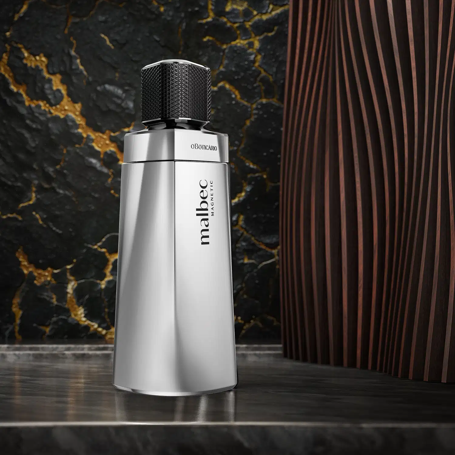 O frasco é prateado e possui um design elegante, com uma tampa preta. O frasco está posicionado em um ambiente com um fundo escuro com veios amarelos e parede com textura amadeirada, sobre superfície de pedra polida cinza.