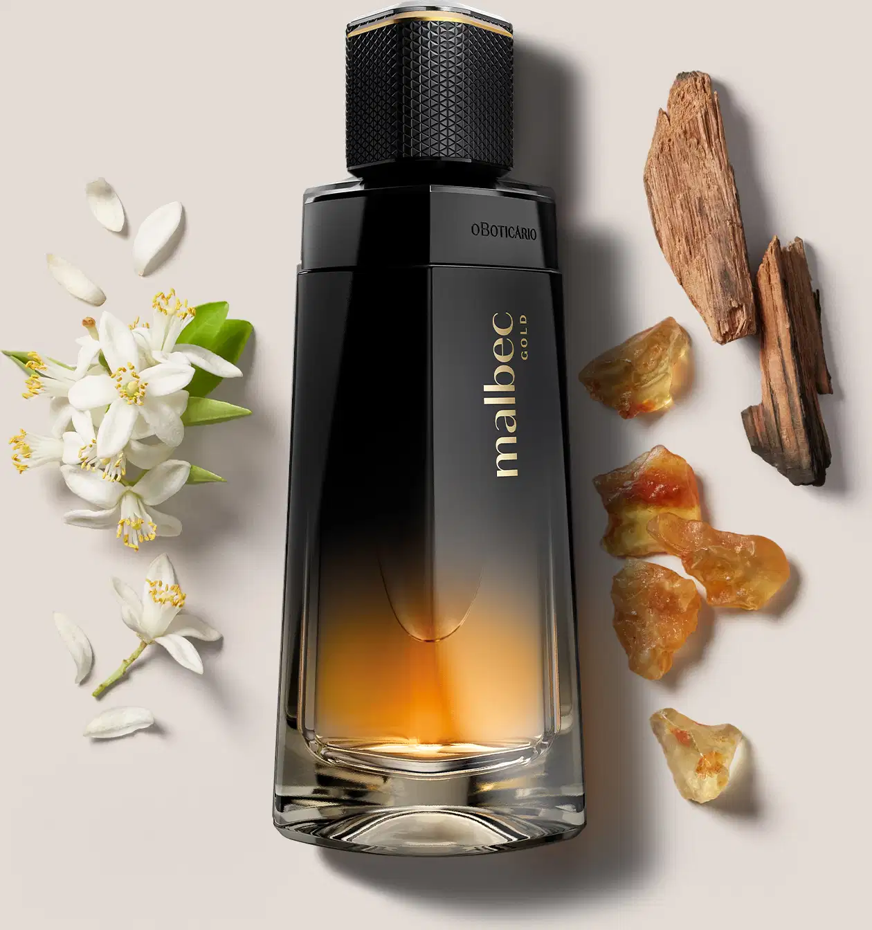 Frasco de desodorante colônia Malbec Gold nas cores degradê de preto com dourado cercado por elementos naturais, como flores brancas e pepitas de âmbar e madeiras, sugerindo ingredientes naturais que compõem a fragrância.