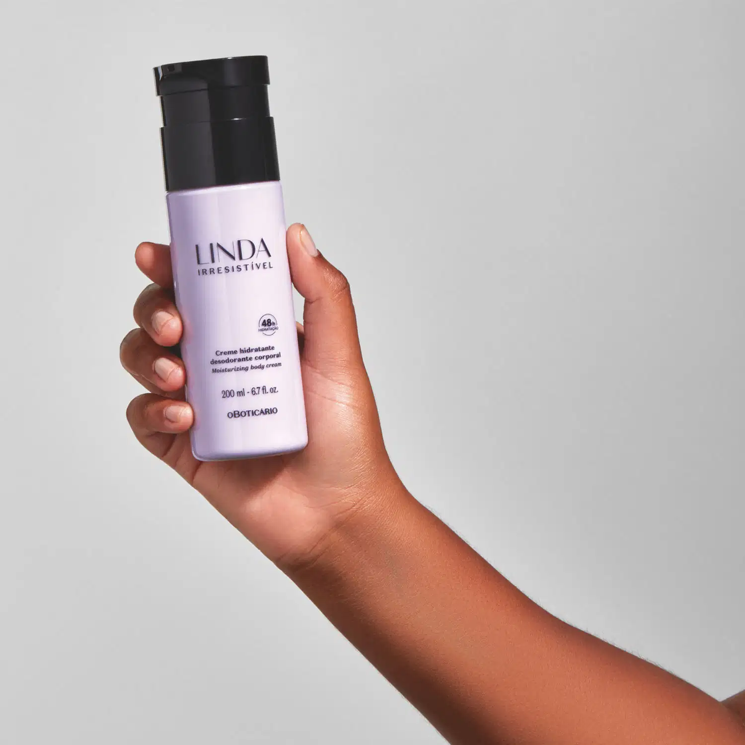 Mão feminina segurando um frasco lilás com tampa e letras pretas de loção hidratante corporal da marca Linda Irresistível em fundo azul claro.