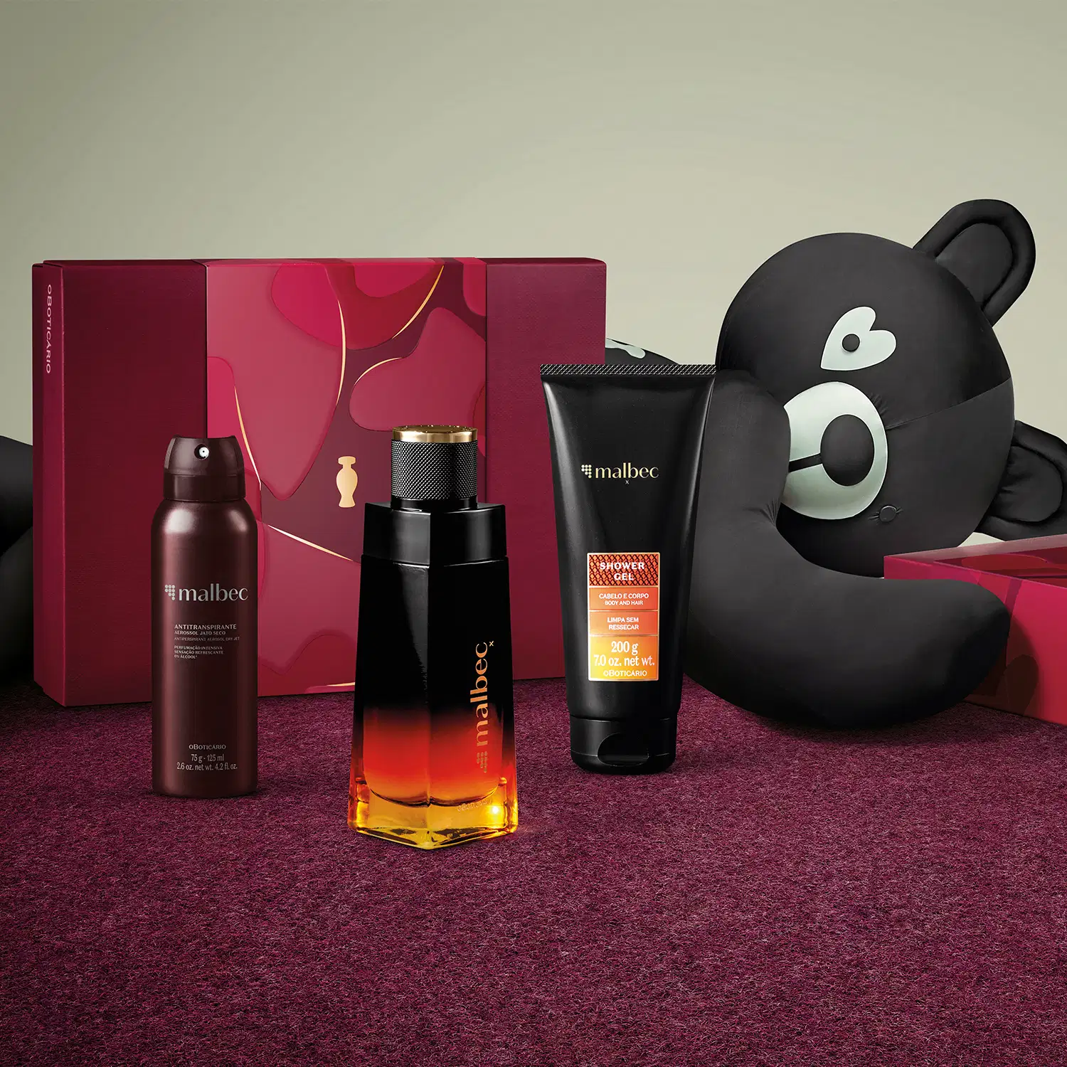 Frasco de desodorante colônia, bisnaga de gel de banho, embalagem de antitranspirante aerossol jato seco e caixa vinho ao lado de um urso de tecido preto e branco em superfície vermelha com floral e fundo cinza.