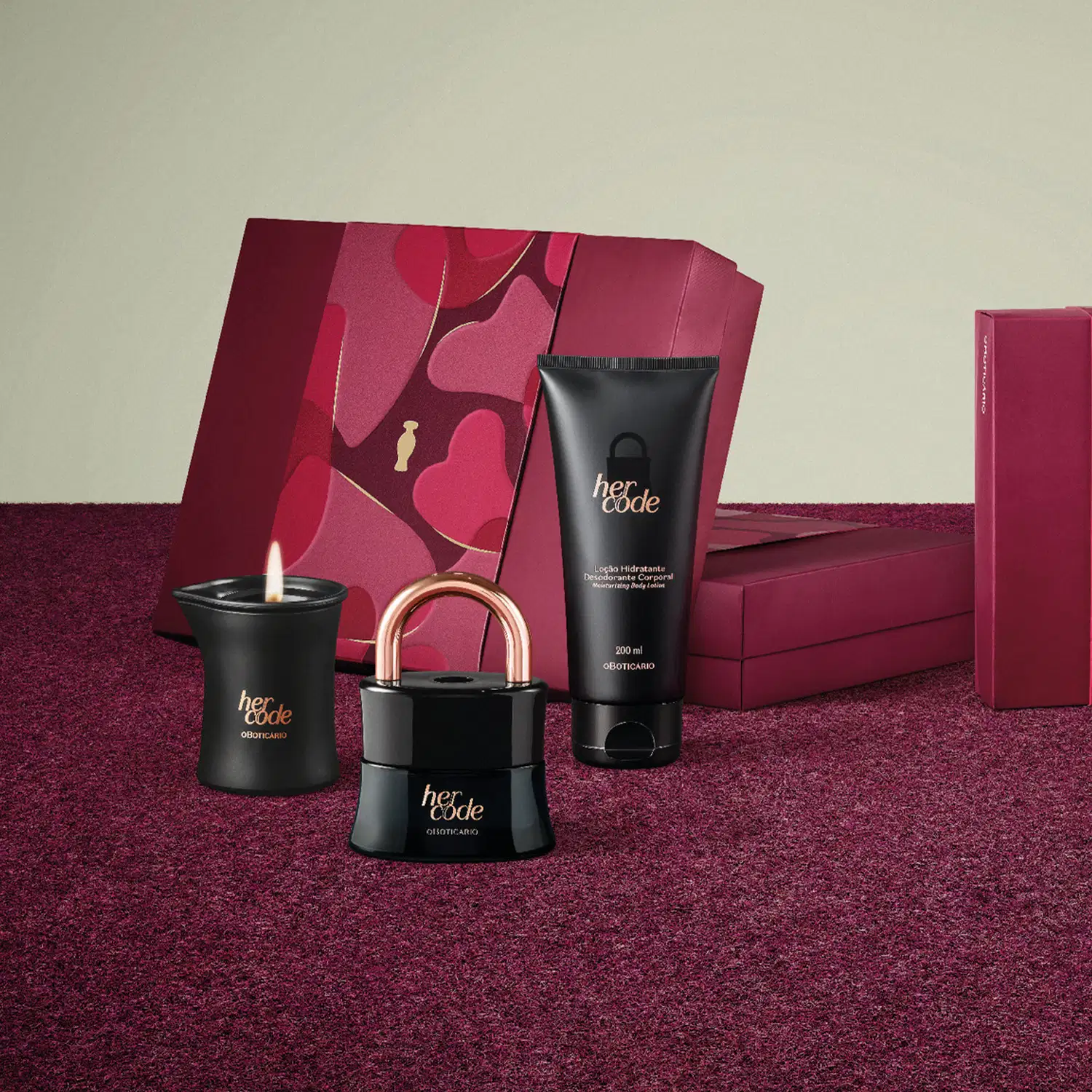 Kit de produtos da marca Her Code, incluindo um frasco de loção hidratante corporal, uma vela aromática e um eau de parfum em formato de cadeado, todos na cor preta e em superfície na cor vinho com detalhe floral e fundo cinza.